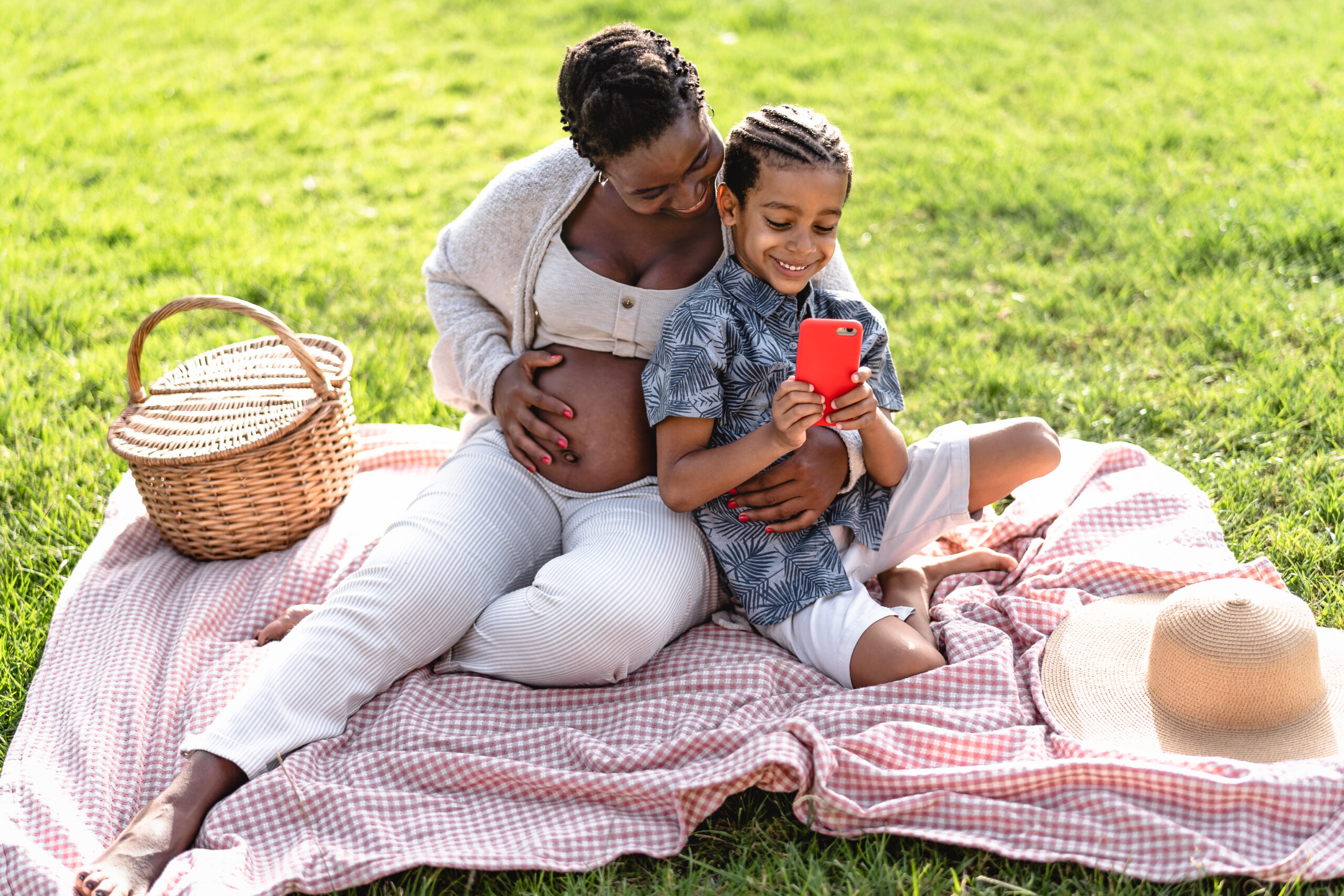 Happy African pregnant mother spending time with his son doing a picnic during weekend in public park - Afro family watching on smartphone - Maternity and parents lifestyle concept